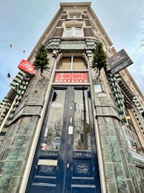 A tall, ornate building facade with an entrance featuring a dark, intricately designed wooden door. The architecture includes green-tiled walls and windows with white decorative elements. Several signs, including one for 'Mosselen', are visible along with small evergreen trees for decoration.