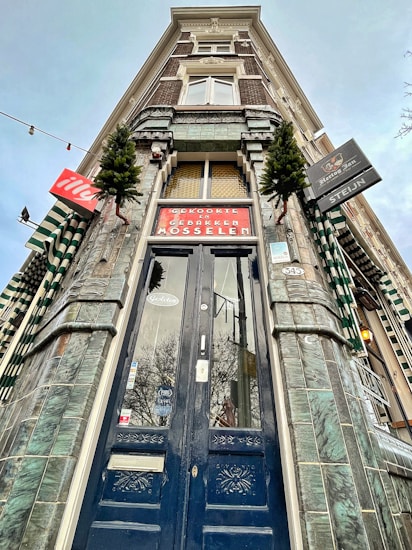 A tall, ornate building facade with an entrance featuring a dark, intricately designed wooden door. The architecture includes green-tiled walls and windows with white decorative elements. Several signs, including one for 'Mosselen', are visible along with small evergreen trees for decoration.