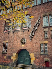 A brick building with a large green door and arched windows. The facade features an artistic installation with the words 'I have to change to stay the same' near the upper right section. Yellow leaves from a tree in the foreground contrast against the brickwork, suggesting a season of change.