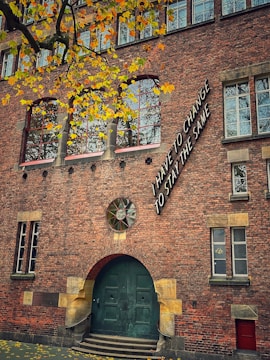 A brick building with a large green door and arched windows. The facade features an artistic installation with the words 'I have to change to stay the same' near the upper right section. Yellow leaves from a tree in the foreground contrast against the brickwork, suggesting a season of change.