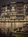 An elegant, multi-story hotel building with illuminated windows and ornate balconies is reflected on a calm body of water. The facade features decorative lighting, creating a warm and inviting atmosphere. A boat adorned with festive garlands is docked in the foreground.