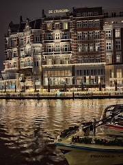 An elegant, multi-story hotel building with illuminated windows and ornate balconies is reflected on a calm body of water. The facade features decorative lighting, creating a warm and inviting atmosphere. A boat adorned with festive garlands is docked in the foreground.