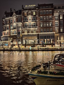 An elegant, multi-story hotel building with illuminated windows and ornate balconies is reflected on a calm body of water. The facade features decorative lighting, creating a warm and inviting atmosphere. A boat adorned with festive garlands is docked in the foreground.