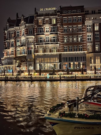 An elegant, multi-story hotel building with illuminated windows and ornate balconies is reflected on a calm body of water. The facade features decorative lighting, creating a warm and inviting atmosphere. A boat adorned with festive garlands is docked in the foreground.