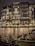 An elegant, multi-story hotel building with illuminated windows and ornate balconies is reflected on a calm body of water. The facade features decorative lighting, creating a warm and inviting atmosphere. A boat adorned with festive garlands is docked in the foreground.