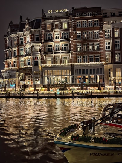 An elegant, multi-story hotel building with illuminated windows and ornate balconies is reflected on a calm body of water. The facade features decorative lighting, creating a warm and inviting atmosphere. A boat adorned with festive garlands is docked in the foreground.