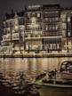 An elegant, multi-story hotel building with illuminated windows and ornate balconies is reflected on a calm body of water. The facade features decorative lighting, creating a warm and inviting atmosphere. A boat adorned with festive garlands is docked in the foreground.