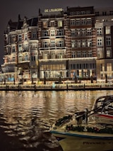 An elegant, multi-story hotel building with illuminated windows and ornate balconies is reflected on a calm body of water. The facade features decorative lighting, creating a warm and inviting atmosphere. A boat adorned with festive garlands is docked in the foreground.