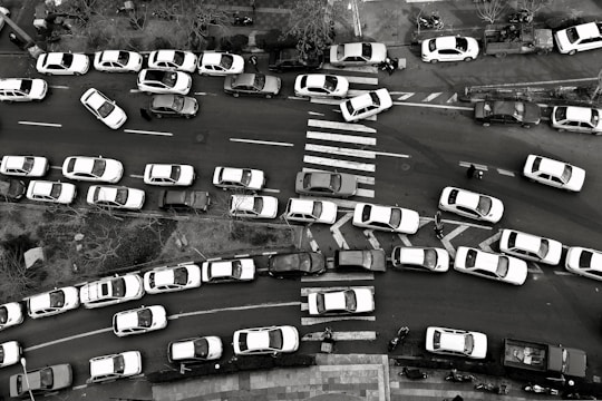 An aerial view captures heavy traffic congestion on a city street. Numerous cars are lined up in multiple lanes with some vehicles attempting to maneuver around others. A zebra crossing is visible, while pedestrians and a few cyclists try to navigate through the packed scene. Trees and sidewalk areas frame the street.