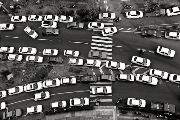 An aerial view captures heavy traffic congestion on a city street. Numerous cars are lined up in multiple lanes with some vehicles attempting to maneuver around others. A zebra crossing is visible, while pedestrians and a few cyclists try to navigate through the packed scene. Trees and sidewalk areas frame the street.