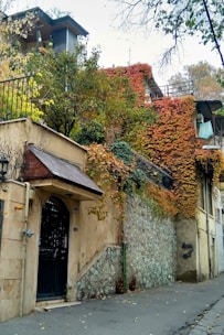 A residential building with an ivy-covered facade, featuring a stone and plaster exterior. The building is surrounded by trees with green, yellow, and orange leaves, indicating an autumn setting. There is a black wrought iron gate at the entrance and a small balcony.