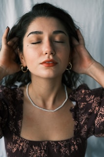 A woman with closed eyes stands in a calm and serene pose. She has long dark hair with a gentle wave, adorned with hoop earrings. Her hands are gently touching the sides of her head. The woman wears a patterned dark blouse and a delicate necklace. The background is a simple light-colored curtain.