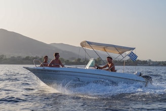 A motorboat with three people is cruising on a body of water. The boat is moving at speed, creating visible splashes and a wake. A Greek flag is attached to the boat. In the background, there are hills and a coastline with trees and buildings under a clear sky.