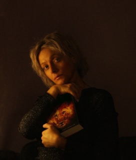 A person with short, light-colored hair is holding a book featuring a lion's face on the cover against a dark background. The lighting creates a warm, intimate atmosphere, highlighting their face and hands.