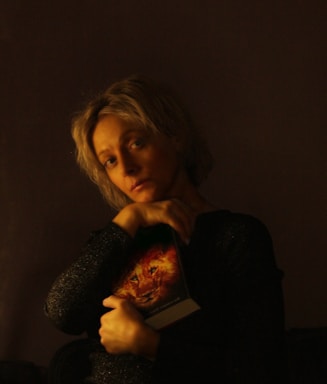 A person with short, light-colored hair is holding a book featuring a lion's face on the cover against a dark background. The lighting creates a warm, intimate atmosphere, highlighting their face and hands.