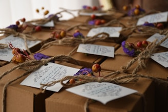 Brown cardboard gift boxes are tied with twine, decorated with small dried flowers and tags containing handwritten messages. The overall presentation is rustic and elegant.