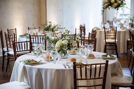 Round tables are elegantly set with white tablecloths and decorated with floral centerpieces. Each table is equipped with glasses, plates, cutlery, and napkins, arranged neatly for a formal event. Wooden chairs encircle the tables in a warm, softly lit environment reminiscent of a wedding or banquet setting.