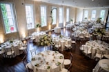 A large, elegantly decorated banquet hall featuring round tables set for dining with white tablecloths and floral centerpieces. Wooden chairs surround each table, and the room is illuminated by natural light coming through tall windows. The space is adorned with greenery and floral arrangements, creating a sophisticated and inviting atmosphere.