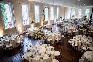 A large, elegantly decorated banquet hall featuring round tables set for dining with white tablecloths and floral centerpieces. Wooden chairs surround each table, and the room is illuminated by natural light coming through tall windows. The space is adorned with greenery and floral arrangements, creating a sophisticated and inviting atmosphere.
