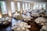 A large, elegantly decorated banquet hall featuring round tables set for dining with white tablecloths and floral centerpieces. Wooden chairs surround each table, and the room is illuminated by natural light coming through tall windows. The space is adorned with greenery and floral arrangements, creating a sophisticated and inviting atmosphere.