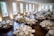 A large, elegantly decorated banquet hall featuring round tables set for dining with white tablecloths and floral centerpieces. Wooden chairs surround each table, and the room is illuminated by natural light coming through tall windows. The space is adorned with greenery and floral arrangements, creating a sophisticated and inviting atmosphere.