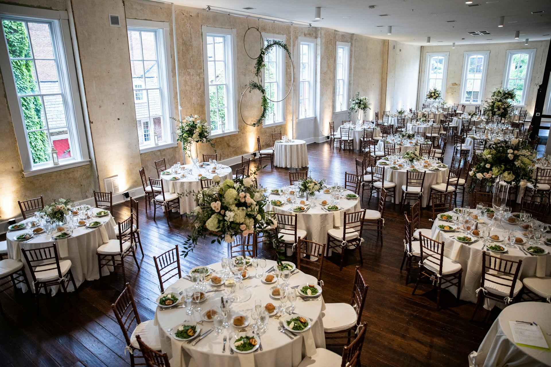 A large, elegantly decorated banquet hall featuring round tables set for dining with white tablecloths and floral centerpieces. Wooden chairs surround each table, and the room is illuminated by natural light coming through tall windows. The space is adorned with greenery and floral arrangements, creating a sophisticated and inviting atmosphere.