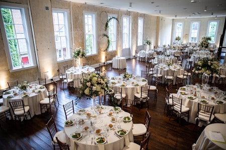 A large, elegantly decorated banquet hall featuring round tables set for dining with white tablecloths and floral centerpieces. Wooden chairs surround each table, and the room is illuminated by natural light coming through tall windows. The space is adorned with greenery and floral arrangements, creating a sophisticated and inviting atmosphere.