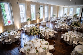 A large, elegantly decorated banquet hall featuring round tables set for dining with white tablecloths and floral centerpieces. Wooden chairs surround each table, and the room is illuminated by natural light coming through tall windows. The space is adorned with greenery and floral arrangements, creating a sophisticated and inviting atmosphere.