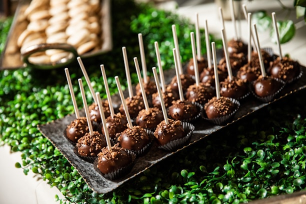 A tray filled with chocolate cake pops, each with a stick inserted, sits atop a bed of green, leafy decorations. In the background, another tray holding unidentified baked goods is partially visible.