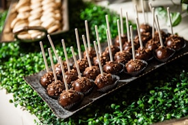 A tray filled with chocolate cake pops, each with a stick inserted, sits atop a bed of green, leafy decorations. In the background, another tray holding unidentified baked goods is partially visible.