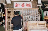 A man stands in front of a metal accordion-style gate, raising a clear plastic curtain to enter a partially open storefront. Above the gate, there is a sign with red Chinese characters. Boxes and various items are visible inside the store, and wooden crates are stacked outside.