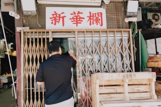 A man stands in front of a metal accordion-style gate, raising a clear plastic curtain to enter a partially open storefront. Above the gate, there is a sign with red Chinese characters. Boxes and various items are visible inside the store, and wooden crates are stacked outside.