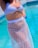 A person is standing beside a pool, wearing a striped bikini top and a sheer, polka dot skirt or cover-up. The water in the pool is a bright blue, giving a refreshing look.