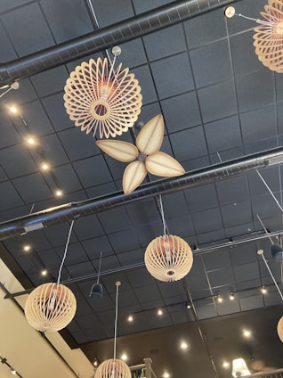 Decorative ceiling installations feature wooden, spherical and petal-like structures suspended by wires, set against a black, grid-tiled ceiling. Small, warmly lit spotlights complement the overall aesthetic.