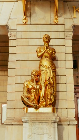 A golden statue featuring two figures: a seated, muscular male figure holding a book, and a standing female figure with hands clasped in front, both set against a stone wall. The artwork is mounted on a pedestal inscribed with the word 'POESIE'.