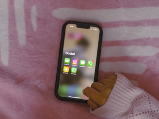 A smartphone displaying a social media folder is being held by a hand wearing a pink sweater. The background consists of a pink and white patterned fabric.