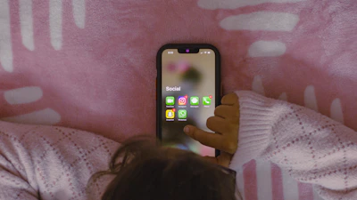 A person is holding a smartphone with several social media applications displayed on the screen. The person is lying on a patterned, pink blanket. The smartphone screen shows apps including Snapchat, Instagram, Facebook Messenger, WhatsApp, and two others.