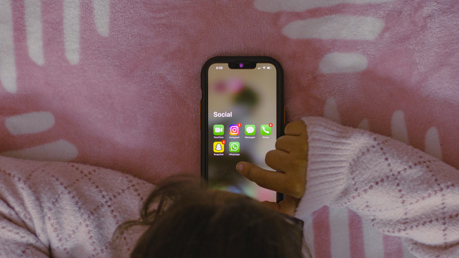 A young child lies in bed seen from behind, reaching up to tap a glowing phone screen showing a 'Social' app folder with Instagram, Snapchat