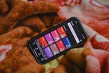A smartphone displaying a music or podcast app screen is placed on a warm, fuzzy blanket. The phone screen shows various categories like 'Telugu,' 'Punjabi,' 'Podcasts,' and others, each highlighted in different colors. The blanket has a soft texture with shades of brown, orange, and red.