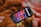 A smartphone displaying a music or podcast app screen is placed on a warm, fuzzy blanket. The phone screen shows various categories like 'Telugu,' 'Punjabi,' 'Podcasts,' and others, each highlighted in different colors. The blanket has a soft texture with shades of brown, orange, and red.