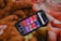 A smartphone displaying a music or podcast app screen is placed on a warm, fuzzy blanket. The phone screen shows various categories like 'Telugu,' 'Punjabi,' 'Podcasts,' and others, each highlighted in different colors. The blanket has a soft texture with shades of brown, orange, and red.