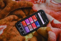 A smartphone displaying a music or podcast app screen is placed on a warm, fuzzy blanket. The phone screen shows various categories like 'Telugu,' 'Punjabi,' 'Podcasts,' and others, each highlighted in different colors. The blanket has a soft texture with shades of brown, orange, and red.