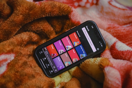 A smartphone displaying a music or podcast app screen is placed on a warm, fuzzy blanket. The phone screen shows various categories like 'Telugu,' 'Punjabi,' 'Podcasts,' and others, each highlighted in different colors. The blanket has a soft texture with shades of brown, orange, and red.