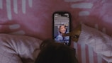 A person wearing a cozy knitted sweater interacts with a smartphone displaying a social media app, against a pink patterned background.