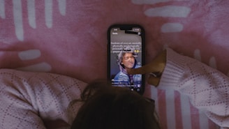 A person wearing a cozy knitted sweater interacts with a smartphone displaying a social media app, against a pink patterned background.
