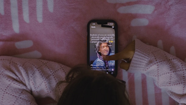 A person wearing a cozy knitted sweater interacts with a smartphone displaying a social media app, against a pink patterned background.