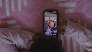 A person wearing a cozy knitted sweater interacts with a smartphone displaying a social media app, against a pink patterned background.
