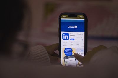 A person is using a smartphone to browse the LinkedIn app in the app store. The screen displays the LinkedIn logo prominently, along with app ratings and download options. The hand holding the phone is partially visible.