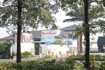 The image features a gated entrance of a modern building with a white facade and dark roof. There are two blue awnings and a distinctive red sign on the building. Surrounding the entrance are various plants and trees. Traffic cones and barriers are positioned near the entrance.