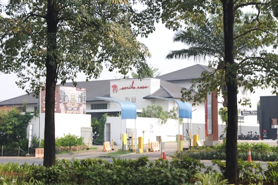 The image features a gated entrance of a modern building with a white facade and dark roof. There are two blue awnings and a distinctive red sign on the building. Surrounding the entrance are various plants and trees. Traffic cones and barriers are positioned near the entrance.
